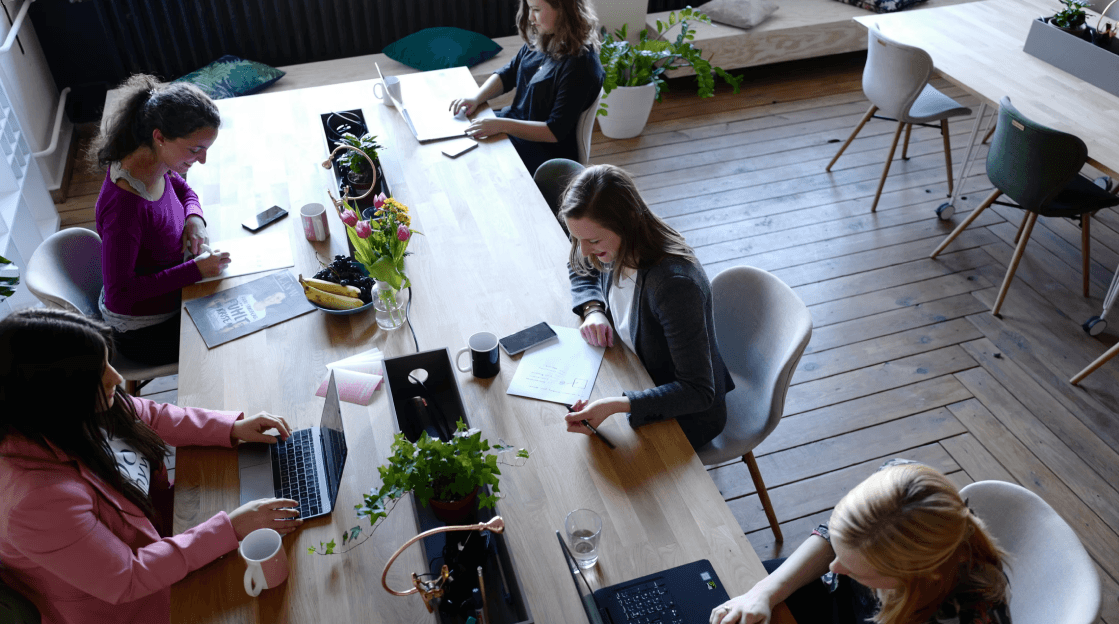 Un groupe de personnes travaillant autour d'une grande table en bois, avec des ordinateurs, des plantes et des tasses, dans un espace lumineux et collaboratif.